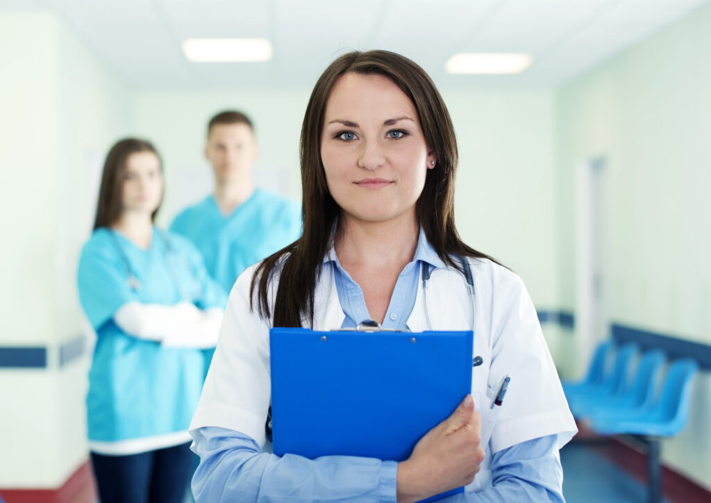 Female doctor holding clipboard in hospital corridor by The Red Pen