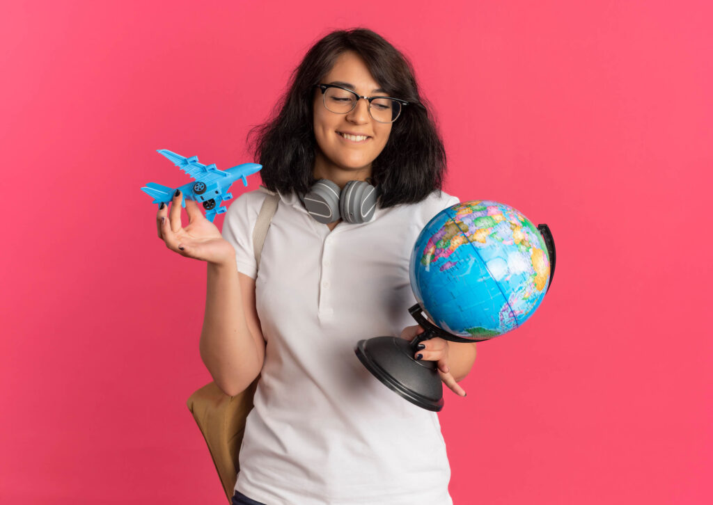 Cheerful female student with headphones, airplane, and globe on pink background, The Red Pen
